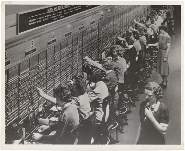 1943年、ベルシステムの国際電話交換台。長い交換台の前に女性交換手が一列に並び、ヘッドセットを着けてケーブルのプラグを抜き差ししている。手前には監督役の女性が立つ