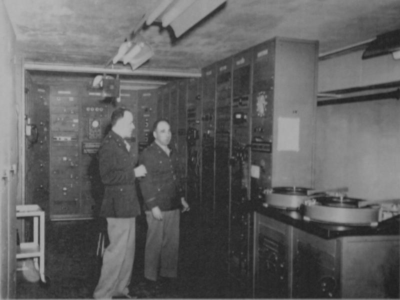 1943年のSIGSALY装置。2人の米陸軍将校が、無数のつまみが並ぶ機器ラックとレコードのターンテーブルの前に立っている